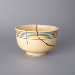 Opposite side view of the Kyoto ware kintsugi cup in cream earthenware, where 24K gold seams branch across the lower body below the blue, green, and gold rim lines.