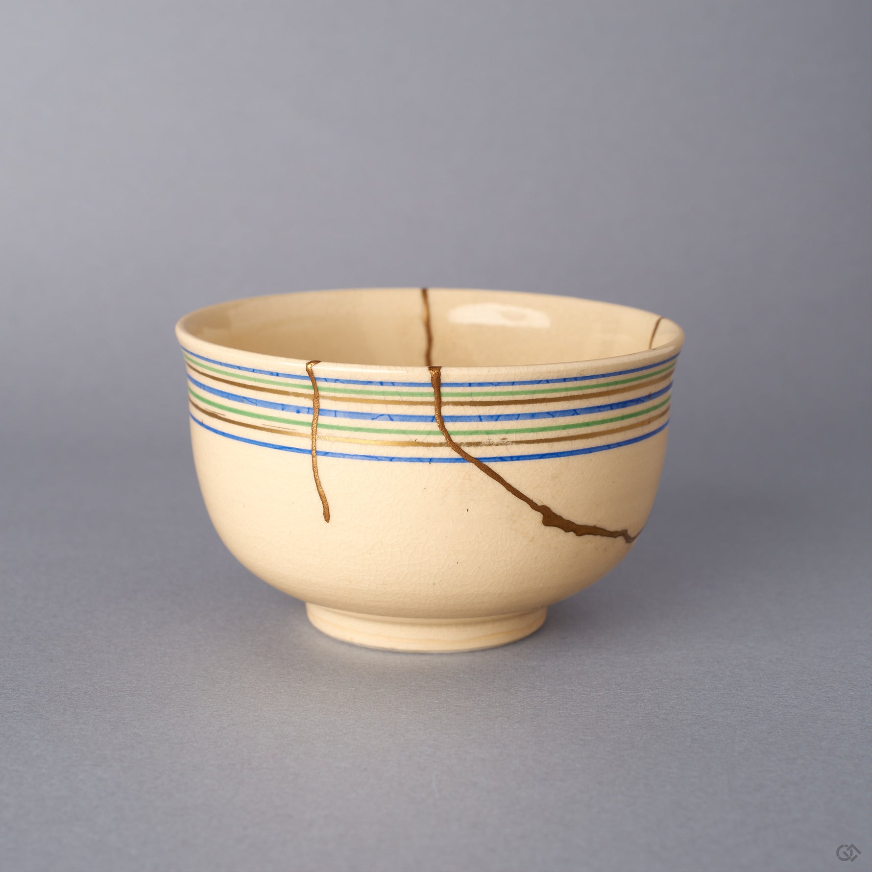 Opposite side view of the Kyoto ware kintsugi cup in cream earthenware, where 24K gold seams branch across the lower body below the blue, green, and gold rim lines.