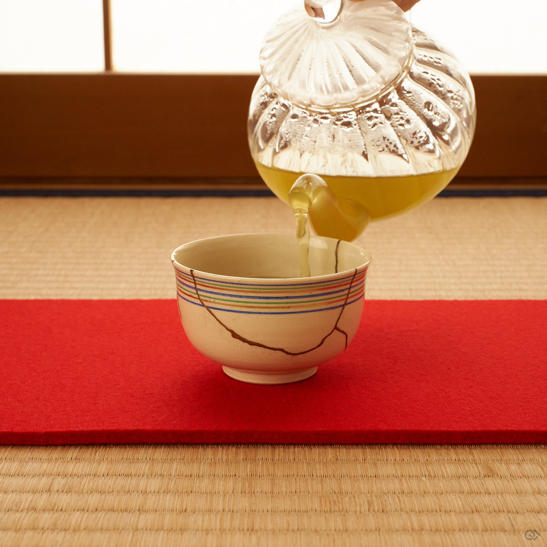 A Kyoto ware kintsugi cup in cream earthenware on a tatami setting, green tea poured from a clear glass teapot, its 24K gold seams catching the light beneath koma-suji lines.