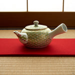 Kintsugi Kyoto ware teapot on a red felt runner over tatami, gold and green shippo pattern across the porcelain body and a nandina branch painted on the restored spout.