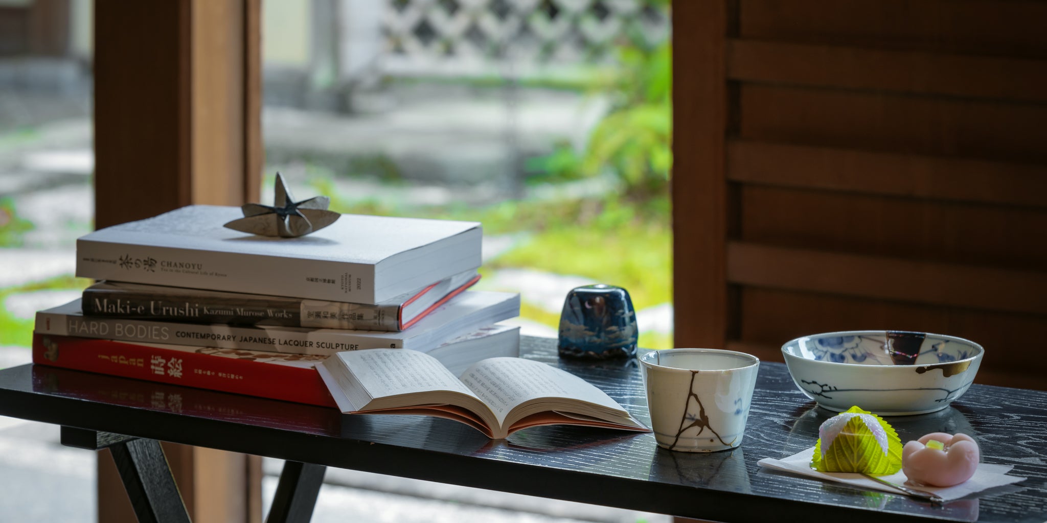 A quiet wabi-sabi moment with Kintsugi art, lacquerware books, wagashi sweets, and The Book of Tea on a black table.