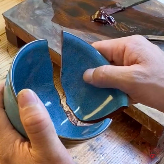 A Japanese artisan restoring a blue matcha bowl with natural urushi lacquer, carefully joining broken ceramic fragments in the traditional Kintsugi method.